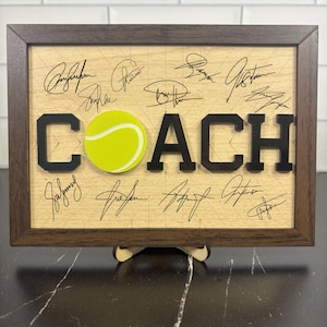 May include: Wooden framed sign with the word "COACH" in black letters, a yellow tennis ball, and signatures. The frame is dark brown, and the background is light wood. A wooden stand supports the sign.