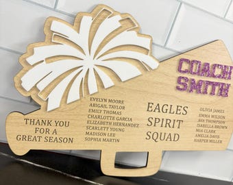 May include: Wooden cheerleading megaphone-shaped plaque with a white pom-pom design. The plaque reads "Coach Smith" in glitter letters, "Eagles Spirit Squad," and "Thank You For A Great Season." Names of cheerleaders are listed.