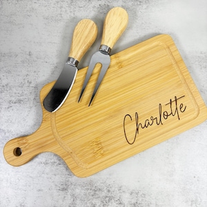 Puede incluir: Una tabla de queso de bambú con un mango, un cuchillo de queso y un tenedor de queso. La tabla está grabada con el nombre "Charlotte". El cuchillo y el tenedor tienen mangos de madera y hojas de acero inoxidable. La tabla es rectangular con esquinas redondeadas.