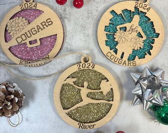 May include: Wooden Christmas ornaments with glitter-filled cutouts. One features a megaphone and the word "Cougars", another a cheerleader silhouette with the name "Lyla", and the third a cheerleader with the name "River". Each ornament has the year "2024".