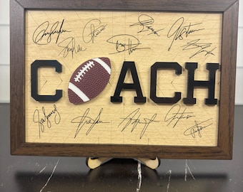 May include: A framed wooden sign with the word "COACH" in black letters and a maroon football in the centre. The sign is filled with signatures and has a dark brown frame. It is displayed on a wooden stand.