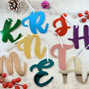 Puede incluir: Adornos de letras de acrílico reflectante de colores, cada uno con un agujero para colgar. Las letras están en una fuente cursiva y se muestran con decoraciones navideñas, incluyendo una piña y bayas rojas.