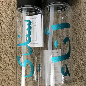 May include: Two clear plastic water bottles with black lids and handles. The bottles have teal Arabic calligraphy on them. One bottle has a sticker with the text "maars" and "24oz Tritan Bottle". The other bottle has a sticker with the text "this product does not include..."