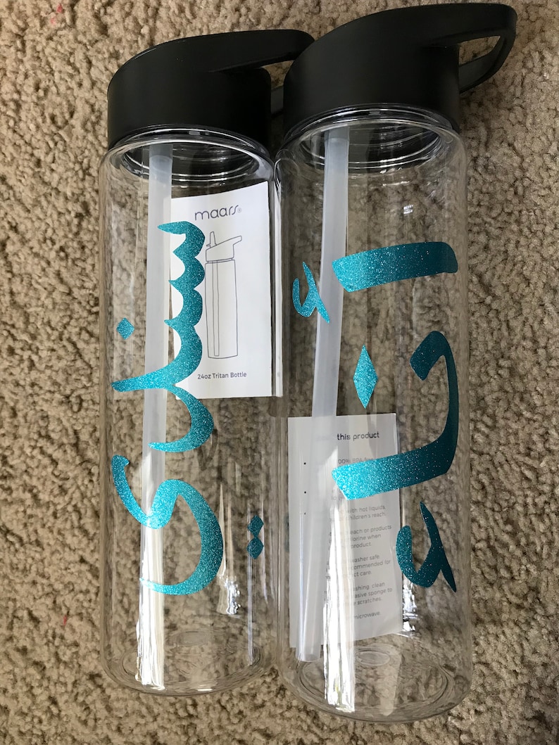 May include: Two clear plastic water bottles with black lids and handles. The bottles have teal Arabic calligraphy on them. One bottle has a sticker with the text "maars" and "24oz Tritan Bottle". The other bottle has a sticker with the text "this product does not include..."