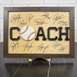 May include: A framed wooden sign with the word "COACH" in black letters and a baseball in the center. The background is light wood with signatures. The frame is dark brown, and the sign is supported by a wooden stand.