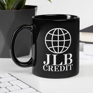Könnte beinhalten: Schwarze Keramik-Tasse mit weißem Globus-Logo und dem Text "JLB CREDIT". Die Tasse hat einen gebogenen Henkel und steht auf einer weißen Oberfläche. Eine Tastatur, eine Pflanze und Bücher sind im Hintergrund.