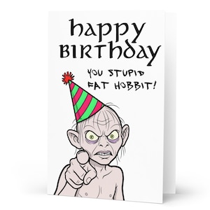 May include: A white greeting card with the words "happy birthday" in a stylized font. The card features a cartoon illustration of Gollum wearing a party hat and pointing, with the text "YOU STUPID FAT HOBBIT!"