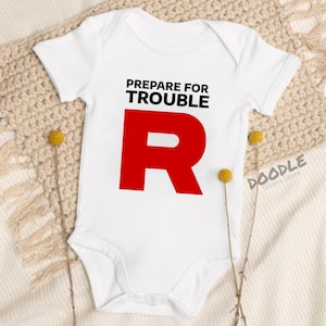 Può includere: Body da bambino bianco con la scritta "PREPARE FOR TROUBLE" in nero sopra una grande lettera "R" rossa. Il body ha maniche corte e chiusura con bottoni a pressione. Il logo Doodle Studio Store è visibile in basso a destra.
