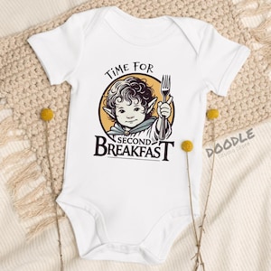 Op de afbeelding: Witte babybody met een afbeelding van een stripfiguur met een vork, met de tekst "TIME FOR SECOND BREAKFAST". Het personage staat in een gele cirkel. Doodle Studio Store logo rechts.