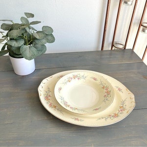 May include: Cream-colored oval serving platter and bowl set with floral and gold trim. The set features a delicate pattern of pink, yellow, and blue flowers. The dishes are placed on a gray wooden surface.