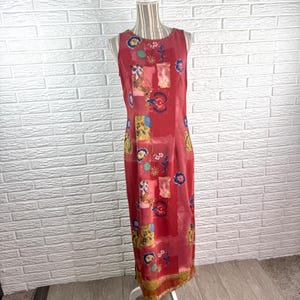 May include: A sleeveless, floor-length dress in a deep red hue, featuring a vibrant, multi-colored floral and abstract pattern. The garment has a round neckline and a straight silhouette, displayed on a mannequin against a white brick wall.
