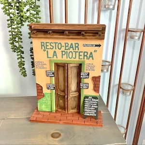 May include: A miniature wooden model of a restaurant called "La Piojera" with a sign above the door that says "Resto-Bar". The model is painted in bright colors and has a brick facade. The restaurant menu is written on a chalkboard.