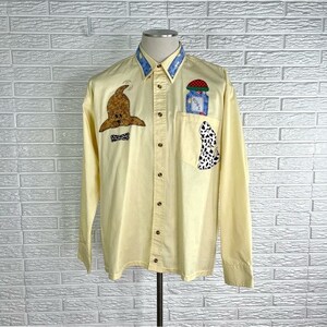 May include: A light yellow button-down shirt with long sleeves. The collar is blue with white stars. The shirt features whimsical patches, including a brown hat, a mushroom, and a black and white spotted design. The shirt is displayed on a mannequin.