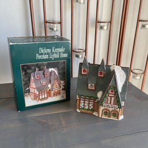 May include: A Dickens Keepsake porcelain lighted house figurine featuring a multi-gabled roof with white snow accents, timber-framed walls, and a sign reading "Welby Gallery". The house is displayed next to its original dark green cardboard box.