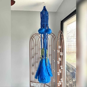 May include: A striking blue macrame plant holder suspended from the ceiling. It features a woven top, two fluffy blue spheres, and long, flowing strands. Greenery adds a decorative touch, making it a unique piece for indoor spaces.