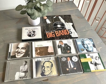 Puede incluir: Una colección de CD de música vintage con artistas como Dino, The Crooners, Al Jolson y Tony Bennett. Los CD se muestran en una superficie de madera con una planta en una maceta blanca en el fondo.
