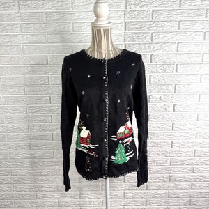 Può includere: Un cardigan nero a maniche lunghe con chiusura a bottoni, decorato con una scena invernale ricamata raffigurante case, alberi di pino verdi e fiocchi di neve bianchi. Il capo presenta uno scollo rotondo e cuciture bianche a vista.