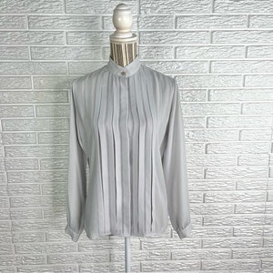 May include: A light gray, long-sleeved blouse with a high collar and vertical pleats. The blouse has a button-up front and is made of a silky fabric.