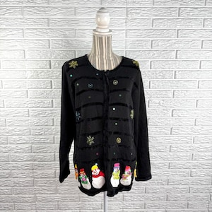 May include: A black long-sleeve cardigan featuring horizontal velvet-like stripes, embroidered gold snowflakes, and colorful sequins. The bottom hem displays a row of embroidered snowmen wearing hats and scarves, with a front button closure.