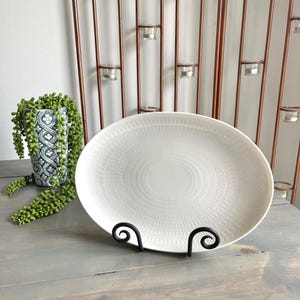 May include: An oval white ceramic serving platter featuring a subtle, embossed radial pattern radiating from the center. The platter is displayed upright on a black metal scrollwork stand, showcasing its textured surface and clean, bright finish.