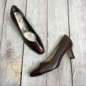 May include: A pair of brown leather pumps with a crocodile-embossed toe. The shoes have a low heel and are labeled "Ross Hommeson".