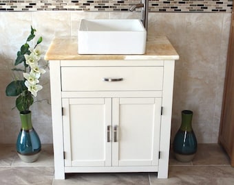 May include: White bathroom vanity cabinet with a white ceramic vessel sink and a chrome faucet. The cabinet has two doors and one drawer. The countertop is made of travertine stone. There are two green and blue vases with flowers on either side of the sink.