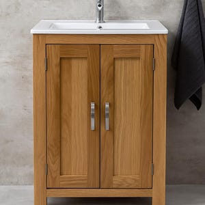 May include: A wooden bathroom vanity unit with a white ceramic sink and a chrome tap. The unit has two doors with silver handles and is made of light-coloured wood. A dark grey towel hangs on the wall to the right.