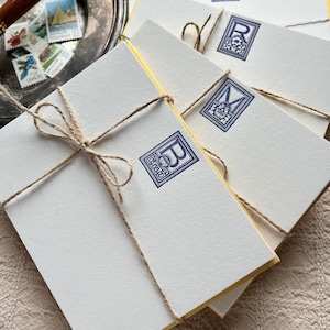 Può includere: Una pila di cancelleria bianca con bordi dorati, legata con spago. Ogni biglietto presenta un timbro blu con monogramma. Un vassoio d'argento con una penna e francobolli sullo sfondo.
