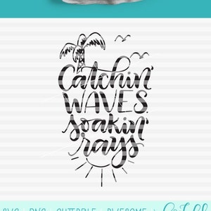 May include: A gray t-shirt with the words "Catchin' Waves, Soakin' Rays" in black script, featuring a palm tree, birds, and sun graphic. The shirt is hanging on a black hanger against a teal background. The bottom graphic repeats the text.