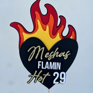 May include: A layered cake topper in the shape of a flame with a black heart. The flame design features red, orange, and yellow layers. The black heart has the text "Meshas FLAMIN Hot 29" in gold glitter and white lettering.