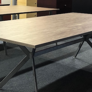 May include: A rectangular office table with a light wood grain surface and a dark gray metal frame. The table has angled legs for stability. The image shows multiple tables in a room.