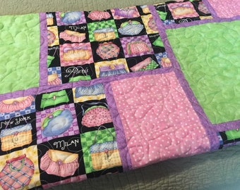Puede incluir: Una manta acolchada con un diseño de patchwork que presenta varios bolsos y las palabras "Nueva York", "Milán" y "París". La manta está hecha con telas verdes, rosas y moradas.