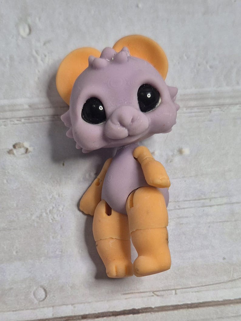 Peut inclure: Une petite souris jouet articul&eacute;e, avec un corps lavande et des oreilles, bras et jambes oranges. Le jouet a de grands yeux noirs et un petit nez rose. Les membres sont articul&eacute;s, permettant le mouvement. Le jouet est con&ccedil;u pour ressembler &agrave; une souris de dessin anim&eacute;.