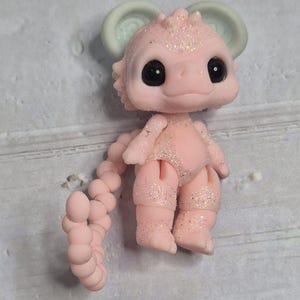 Peut inclure: Figurine rose et paillet&eacute;e d'une cr&eacute;ature fantastique avec de grands yeux noirs, un petit nez et de grandes oreilles. La cr&eacute;ature a une queue segment&eacute;e et est en position verticale. La figurine est sur une surface grise.