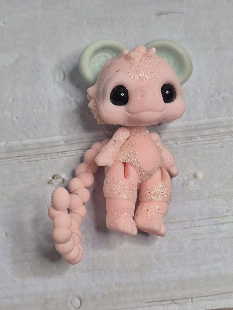 Peut inclure: Figurine rose et paillet&eacute;e d'une cr&eacute;ature fantastique avec de grands yeux noirs, un petit nez et de grandes oreilles. La cr&eacute;ature a une queue segment&eacute;e et est en position verticale. La figurine est sur une surface grise.