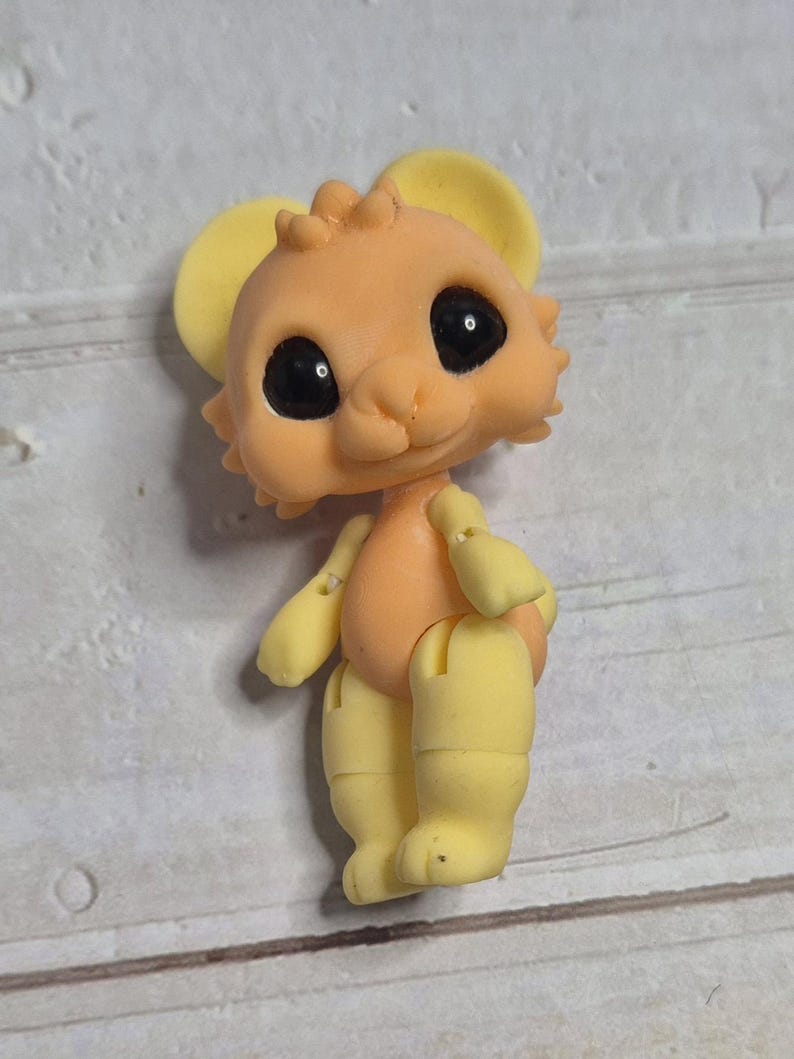 Peut inclure: Petite figurine articul&eacute;e repr&eacute;sentant une souris de dessin anim&eacute;. La souris a un corps couleur p&ecirc;che, des oreilles, des bras et des jambes jaunes, et de grands yeux noirs. La figurine est debout.
