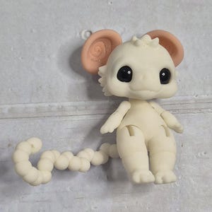 Peut inclure: Petite figurine blanc cass&eacute; repr&eacute;sentant une cr&eacute;ature avec de grands yeux noirs ronds et de grandes oreilles roses claires. La figurine a une queue segment&eacute;e et des membres articul&eacute;s. La cr&eacute;ature est pos&eacute;e sur une surface gris clair.