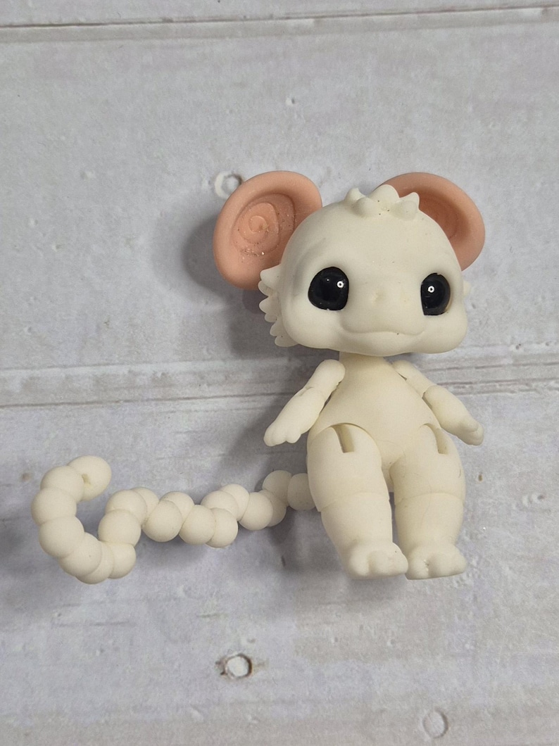 Peut inclure: Petite figurine blanc cass&eacute; repr&eacute;sentant une cr&eacute;ature avec de grands yeux noirs ronds et de grandes oreilles roses claires. La figurine a une queue segment&eacute;e et des membres articul&eacute;s. La cr&eacute;ature est pos&eacute;e sur une surface gris clair.