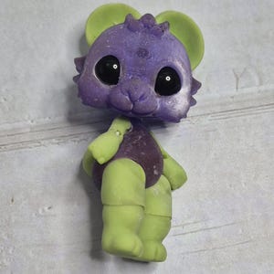 Peut inclure: Figurine fantaisiste d'une cr&eacute;ature avec une t&ecirc;te violette, des oreilles vertes et des membres verts. La cr&eacute;ature a de grands yeux noirs et un corps violet. La figurine est debout, mettant en valeur son design et sa palette de couleurs uniques.