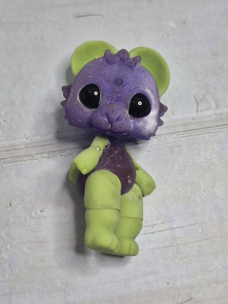 Peut inclure: Figurine fantaisiste d'une cr&eacute;ature avec une t&ecirc;te violette, des oreilles vertes et des membres verts. La cr&eacute;ature a de grands yeux noirs et un corps violet. La figurine est debout, mettant en valeur son design et sa palette de couleurs uniques.