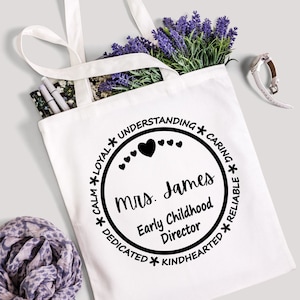 May include: White tote bag with a black circle design. The circle has the text "*LOYAL* UNDERSTANDING *CARING*" around the outside. Inside the circle is the text "mrs. James Early Childhood Director" and a row of small hearts.