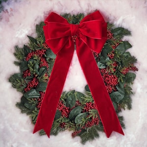 Op de afbeelding: Een rode fluwelen strik zit bovenop een groene krans versierd met rode bessen. De krans is gemaakt van kunstgroen en is perfect voor een feestelijke of winterse decoratie.