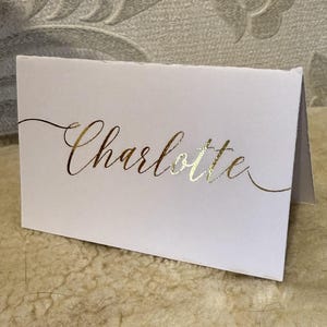 May include: White card with the name "Charlotte" written in gold foil lettering.
