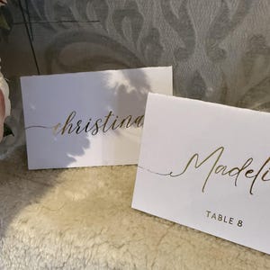 May include: Two white place cards with gold calligraphy. The top card reads "christina" and the bottom card reads "Madeline" with "TABLE 8" below the name.