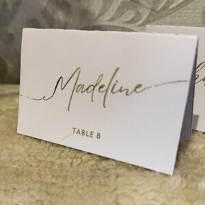 May include: White place card with gold foil lettering. The name "Madeline" is written in cursive script. The text "Table 8" is printed in gold foil below the name.