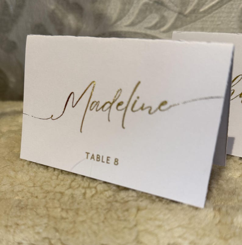 May include: White place card with gold foil lettering. The name "Madeline" is written in cursive script. The text "Table 8" is printed in gold foil below the name.