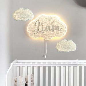 Könnte beinhalten: Eine wolkenförmige Wandleuchte mit dem Namen "Liam" in Schreibschrift. Die Leuchte ist von zwei kleineren wolkenförmigen Dekorationen umgeben. Das Licht ist weiß und hat ein weiches, strukturiertes Aussehen, wobei ein warmes Leuchten von hinter der Wolke ausgeht.