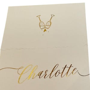 May include: A white place card with gold foil lettering. The text reads "Style 1" at the top and "Charlotte" at the bottom. A gold foil illustration of two champagne glasses clinking together is in the center of the card.