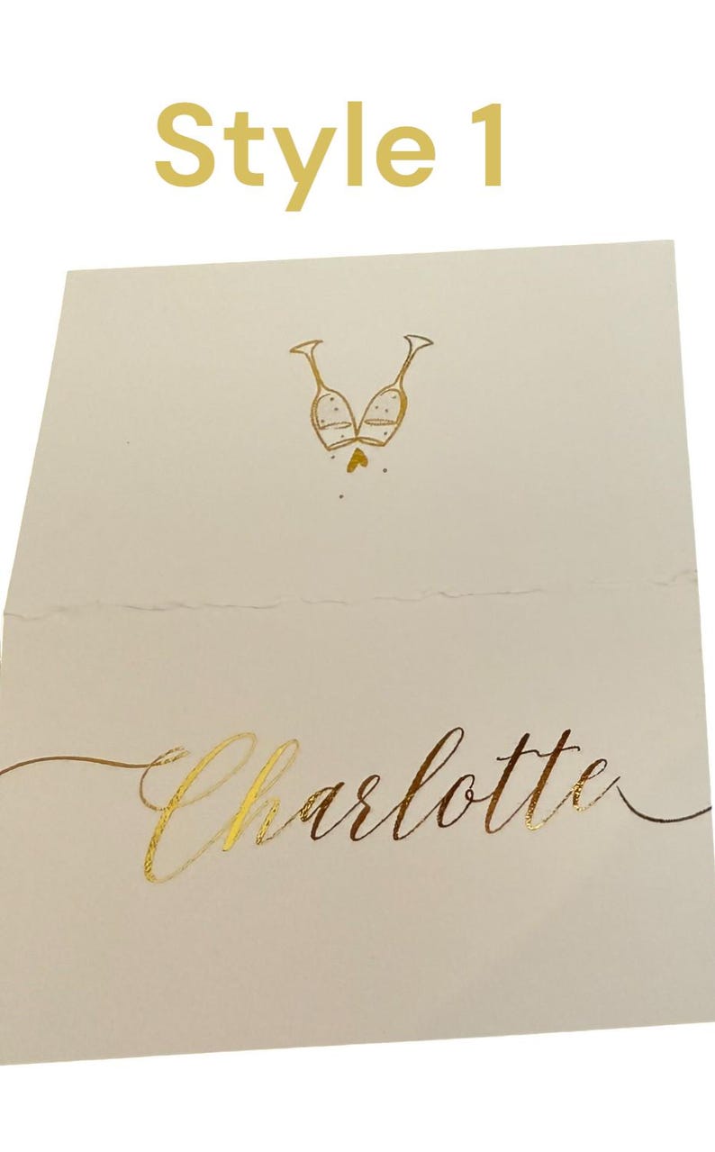 May include: A white place card with gold foil lettering. The text reads "Style 1" at the top and "Charlotte" at the bottom. A gold foil illustration of two champagne glasses clinking together is in the center of the card.