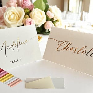 May include: Two white place cards with gold and copper foil lettering. The cards read "Madeline, Table 8" and "Charlotte".